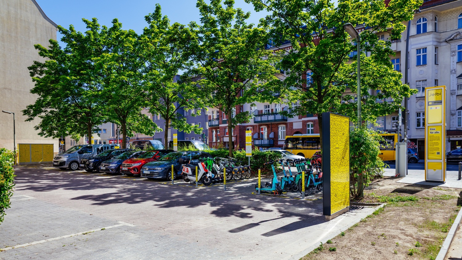 Neben einem Parkplatz mit Autos, Rollern, Scootern und Fahrrädern steht eine Infosäule mit der Überschrift Jelbi.