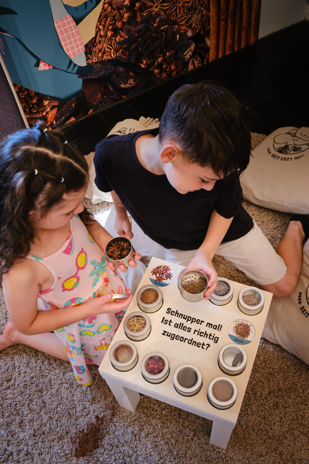 Zwei Kinder hocken und sitzen vor einem kleinen Tisch, auf dem Dosen mit Gewürzen stehen. Auf dem Tisch steht geschrieben "Schnupper mal. Ist alles richtig zugeordnet?".