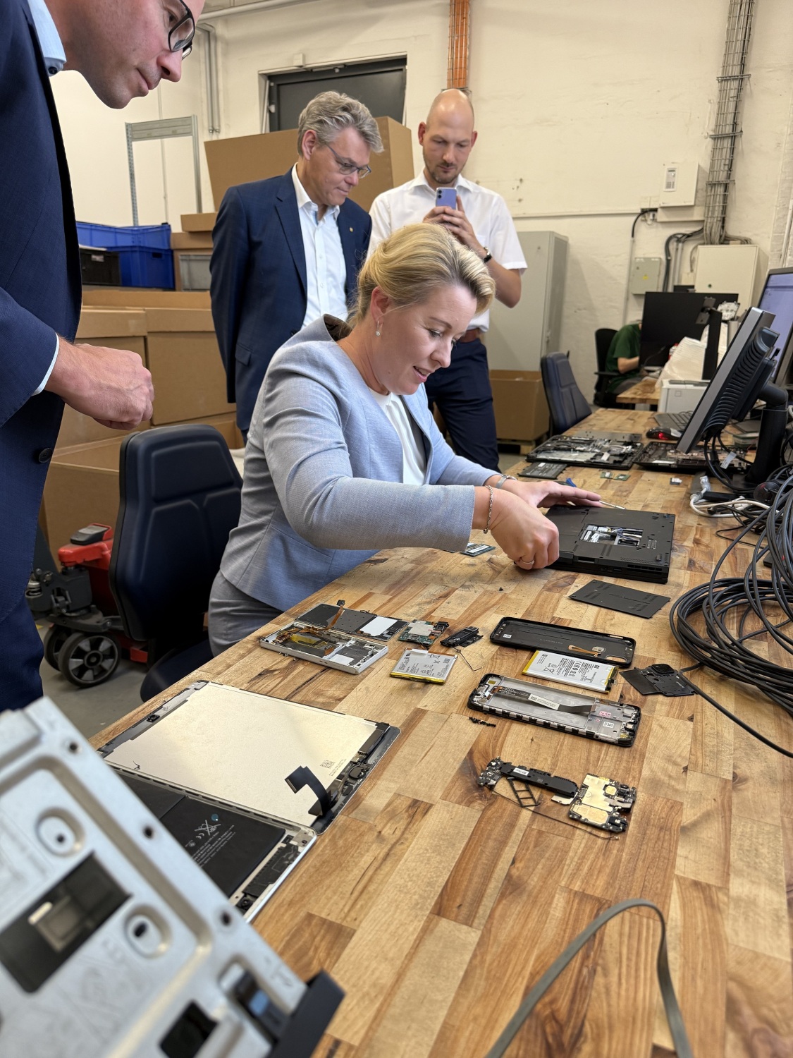 Eine Frau sitzt an einem Tisch mit IT und drei Männer stehen darum und schauen zu.