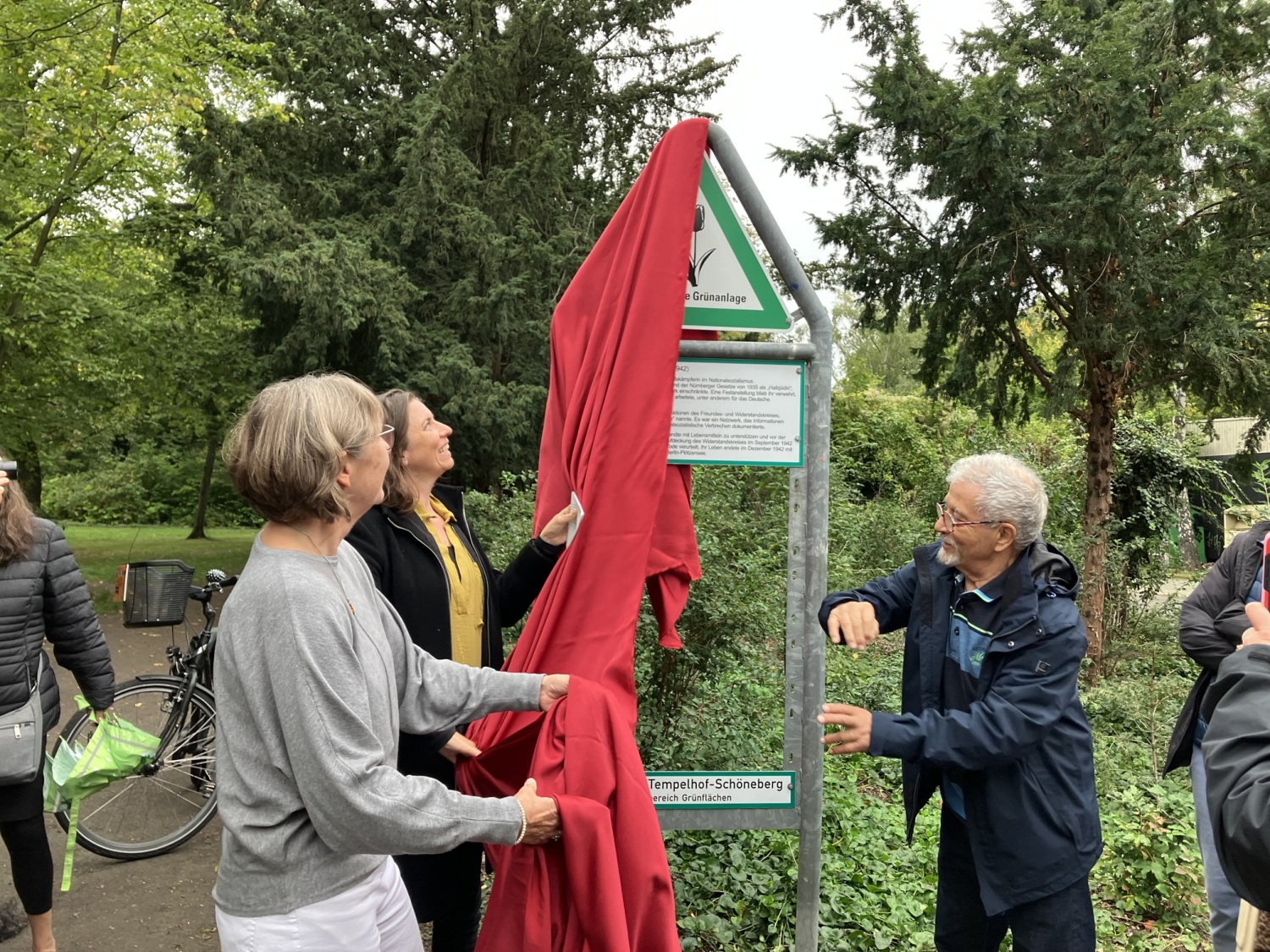 Drei Personen ziehen zusammen ein rotes Tuch von einem Schild am Eingang einer Grünfläche.