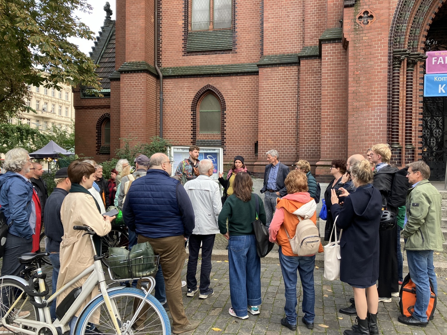 Drei Personen stehen vor einer Menschenmenge und sprechen. Hinter den Personen ist ein großes Backsteingebäude.