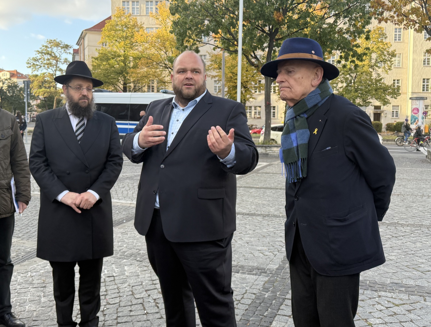 Drei Männer stehen nebeneinander auf einem Platz. Der Mann in der Mitte spricht.