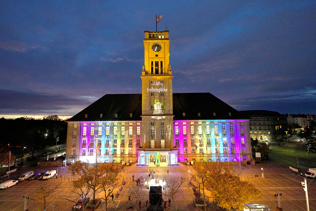 Ein großes Gebäude mit Glockenturm ist nachts bunt beleuchtet, unter anderem mit dem Schriftzug 75 Jahre Freiheitsglocke.