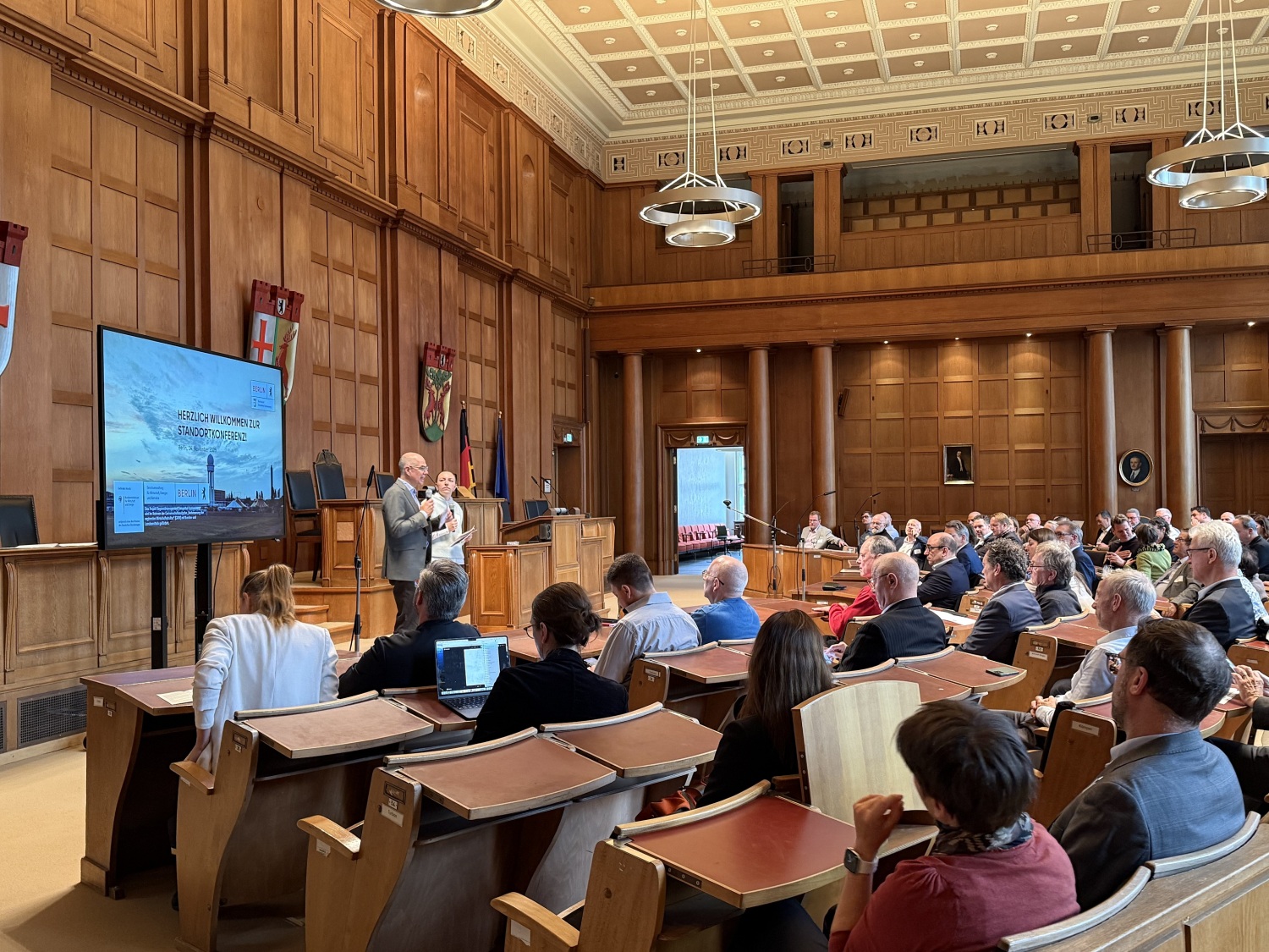 Viele Personen sitzen in Reihen in einem Saal. Vor dem Publikum stehen zwei Menschen vor einem Smartboard und sprechen.