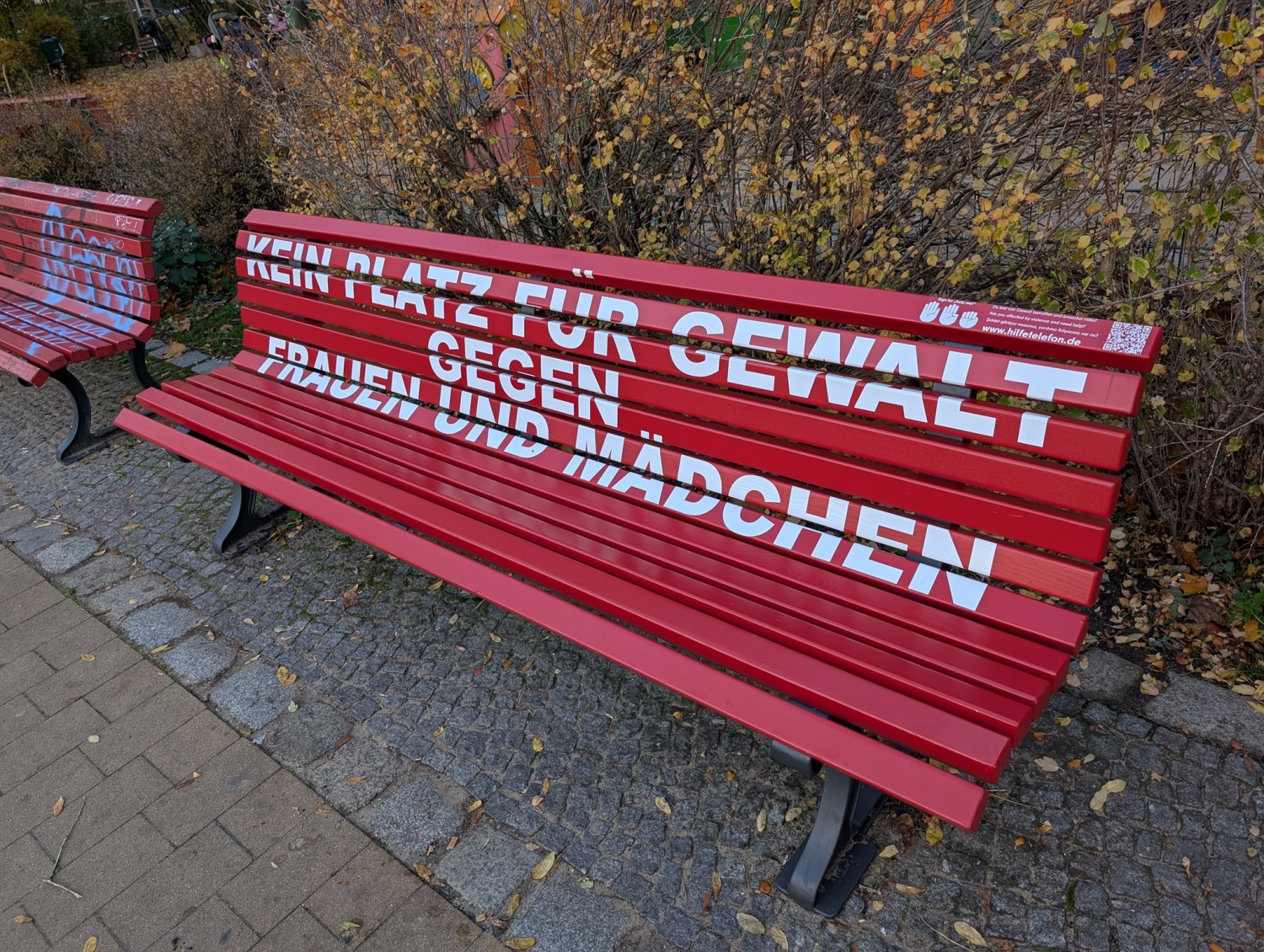 Eine rote Bank mit dem Schriftzug "Kein Platz für Gewalt gegen Frauen und Mädchen". Oben rechts auf der Bank ist ein QR-Code, ein kleiner Text und die Abbildung von Händen.