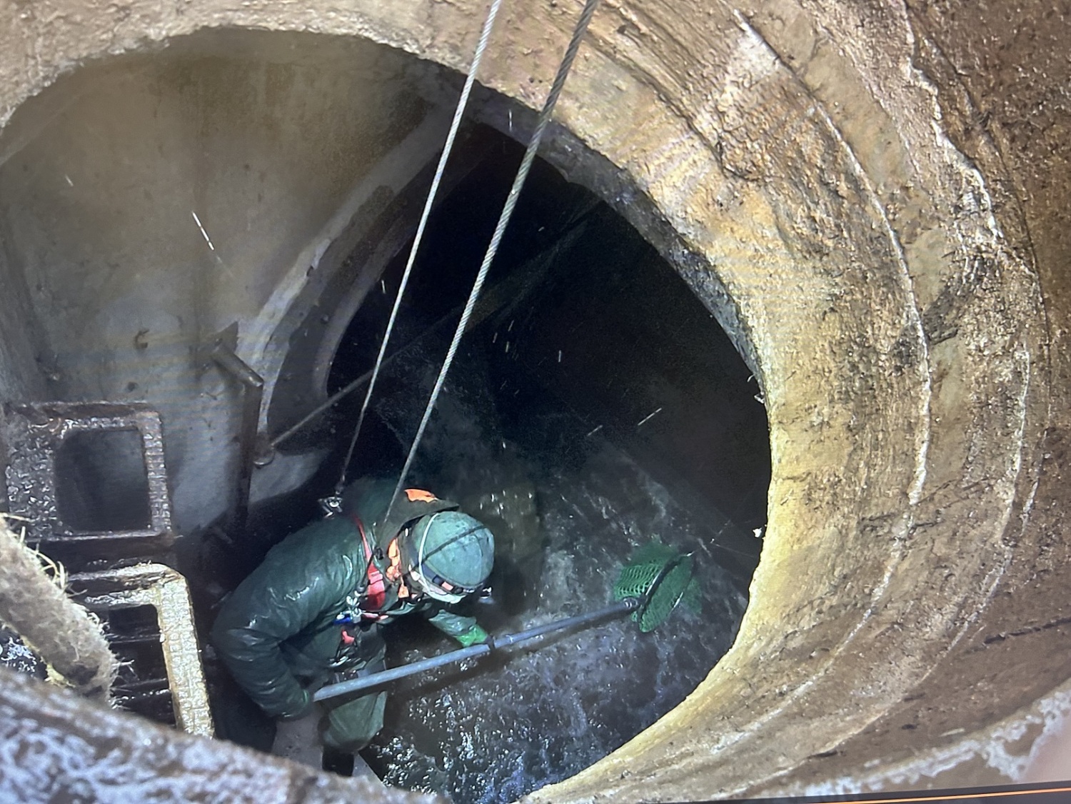 Eine Person mit Spezialkleidung und einem Netz wird durch ein Loch in ein unterirdisches Wasser gelassen.