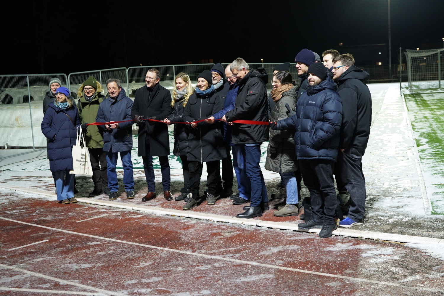 Viele Personen stehen nebeneinander auf einem Sportplatz. Mehrere halten ein rotes Band, was durchschnitten wird.