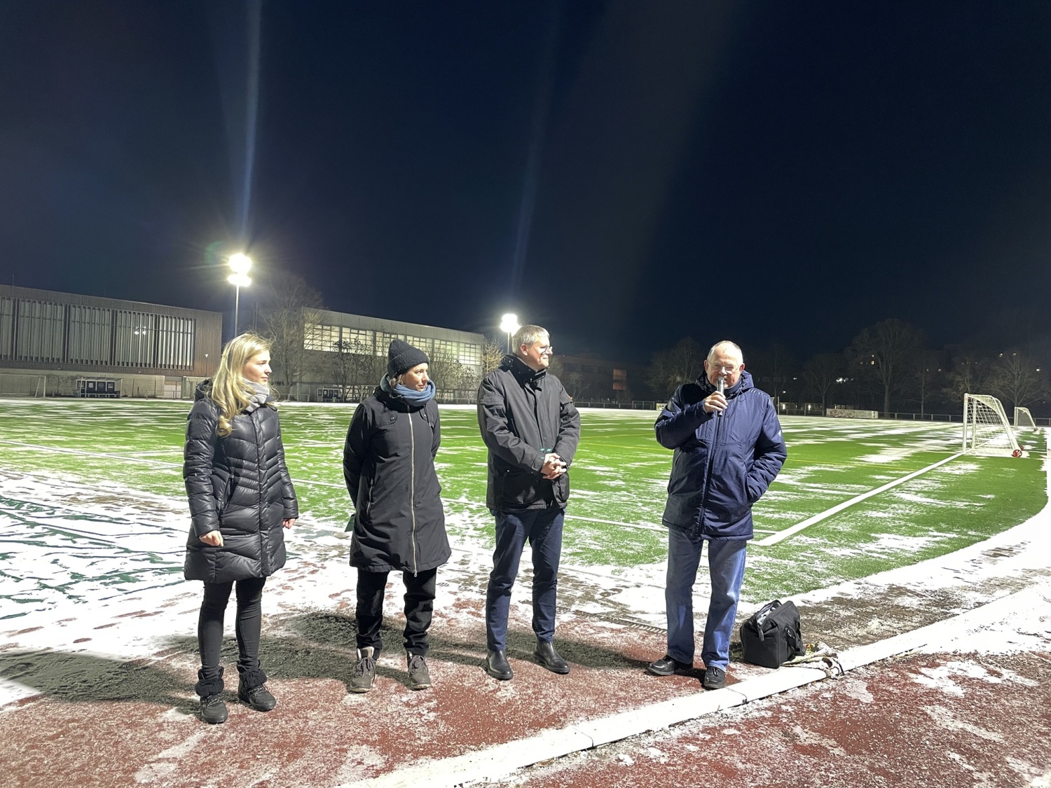 Vier Personen stehen nebeneinander auf einem Sportplatz. Eine Person spricht in ein Mikrofon.