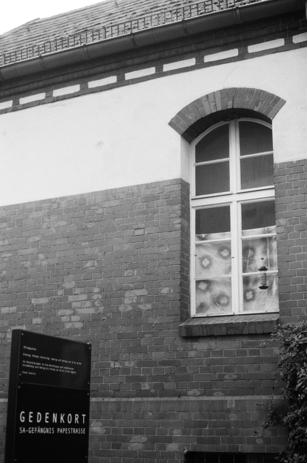 ein schwarz-weiß Foto von einer Hauswand mit einem Fenster, davor ein Schild mit "Gedenkort SA-Gefängnis Papestraße"
