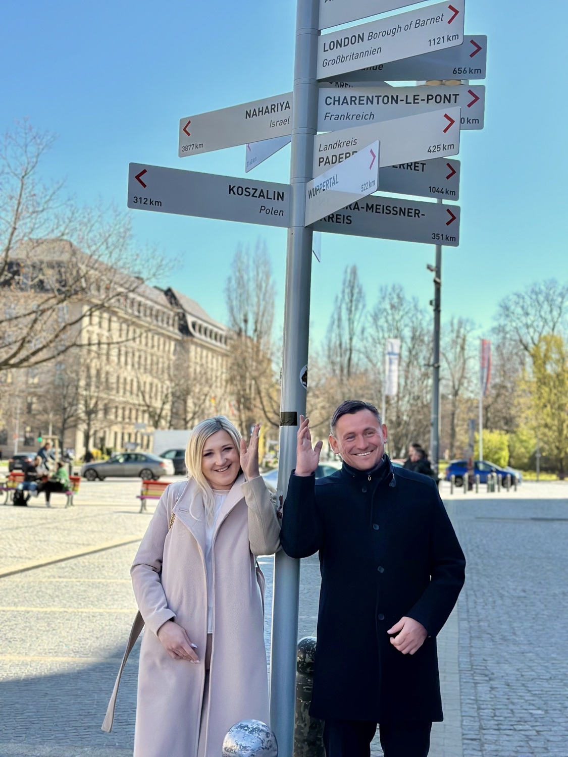 Zwei Personen stehen vor einem Schild, an dem verschiedene Orte mit einer Richtung und Distanz aufgeführt sind.