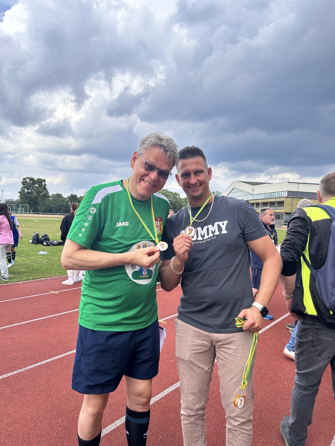 Zwei Männer stehen auf einem Sportplatz nebeneinander und präsentieren ihre Medaillen.