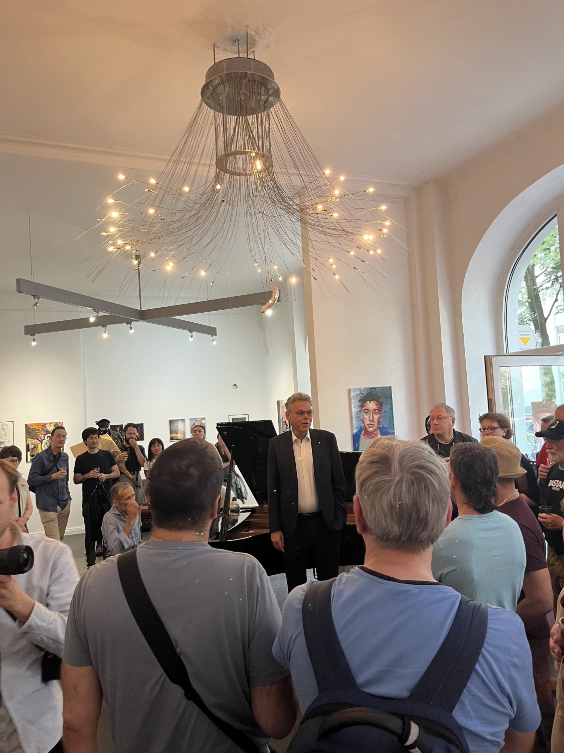Ein Mann steht in der Mitte eines Raumes mit vielen Menschen. Über ihm ist ein Kronleuchter, hinter ihm ist ein Klavier.