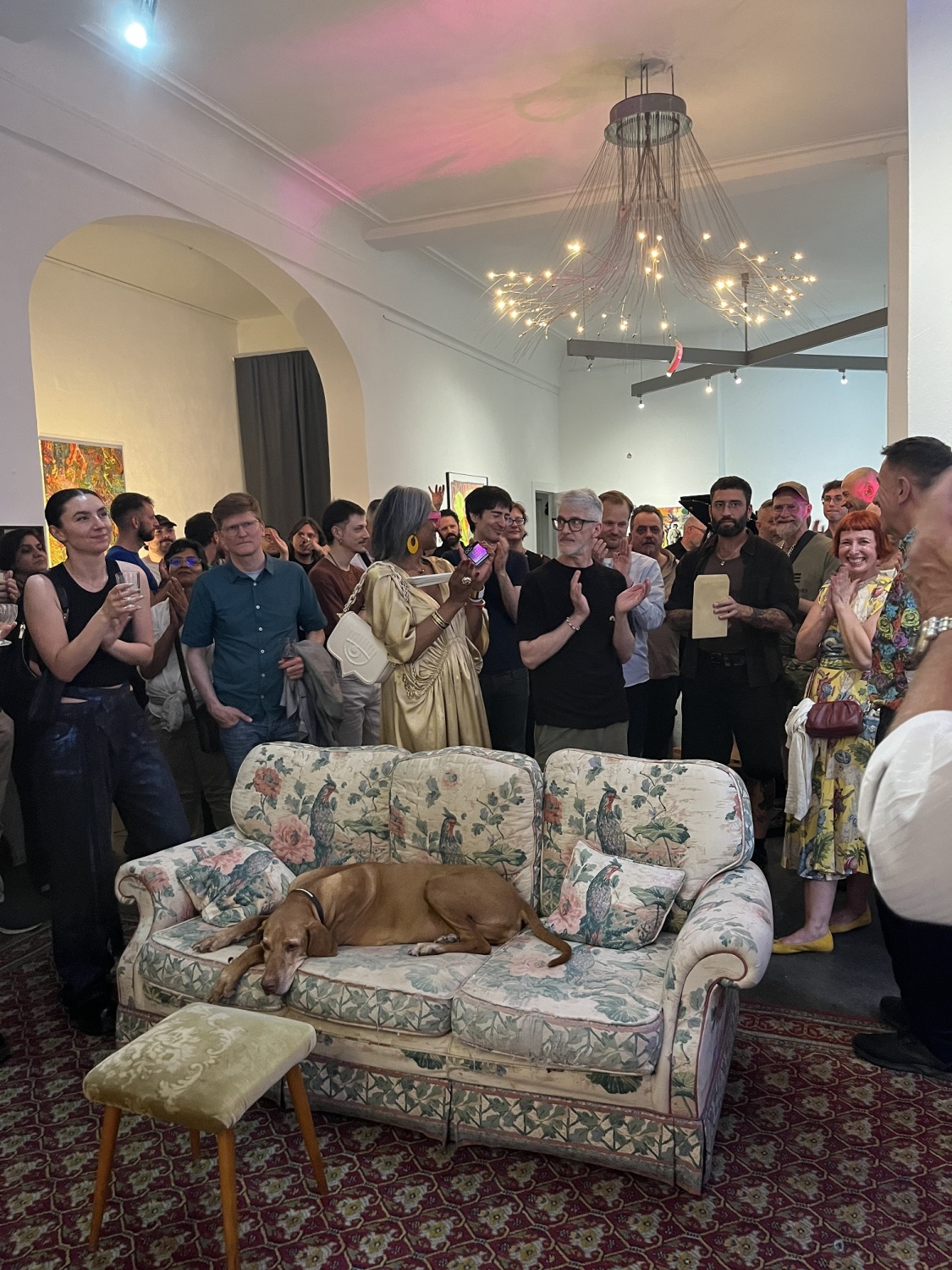 Viele Menschen stehen in einem Raum. Im Vordergrund liegt ein Hund auf einem kleinen Sofa.