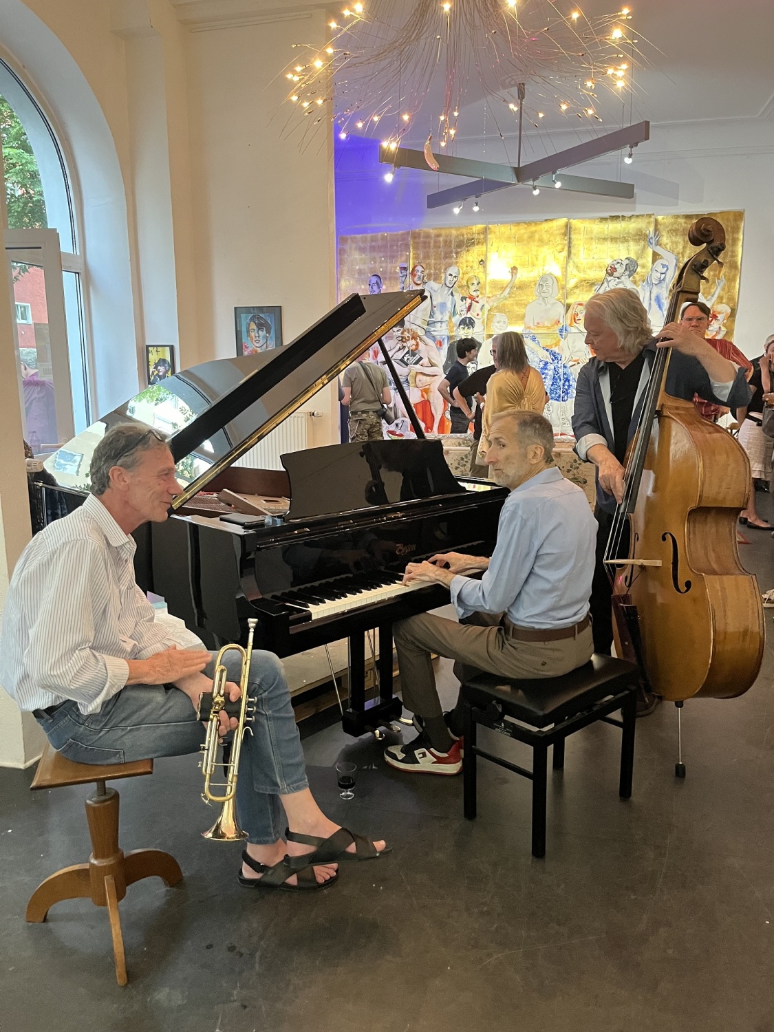 Ein Mann sitzt an einem Klavier, einer sitzt daneben und hat eine Trompete in der Hand und noch ein weiterer Mann hält ein Kontrabass.