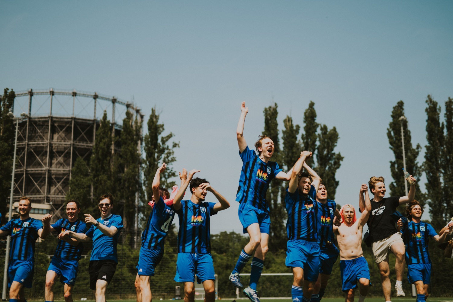 Ein Fußballteam feiert zusammen auf einem Platz. In der Mitte springt jemand in die Luft. Hinter dem Platz ragt ein Gasometer in die Höhe.
