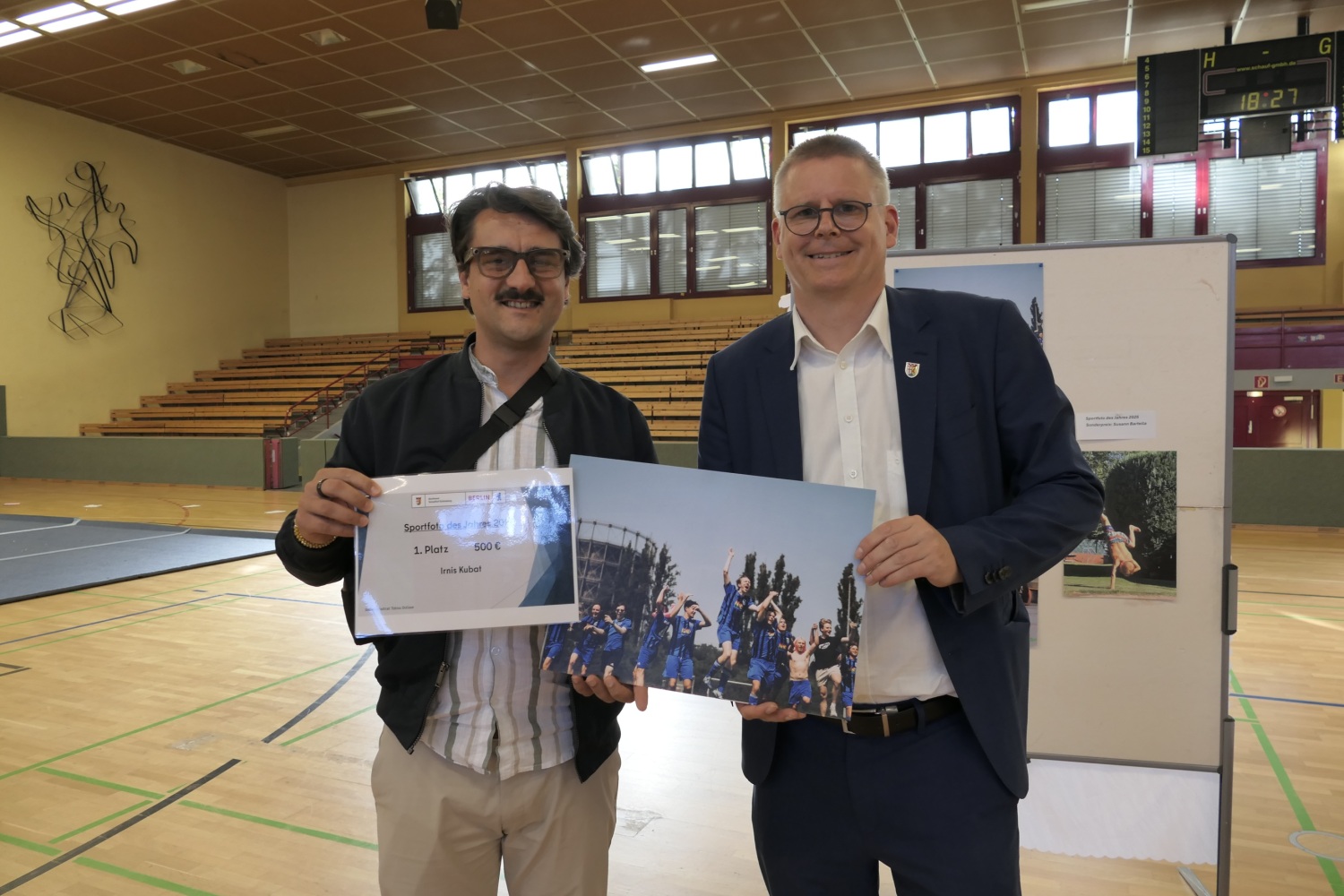 In einer Halle stehen zwei Männer. Einer hält eine Urkunde, der andere ein Foto.