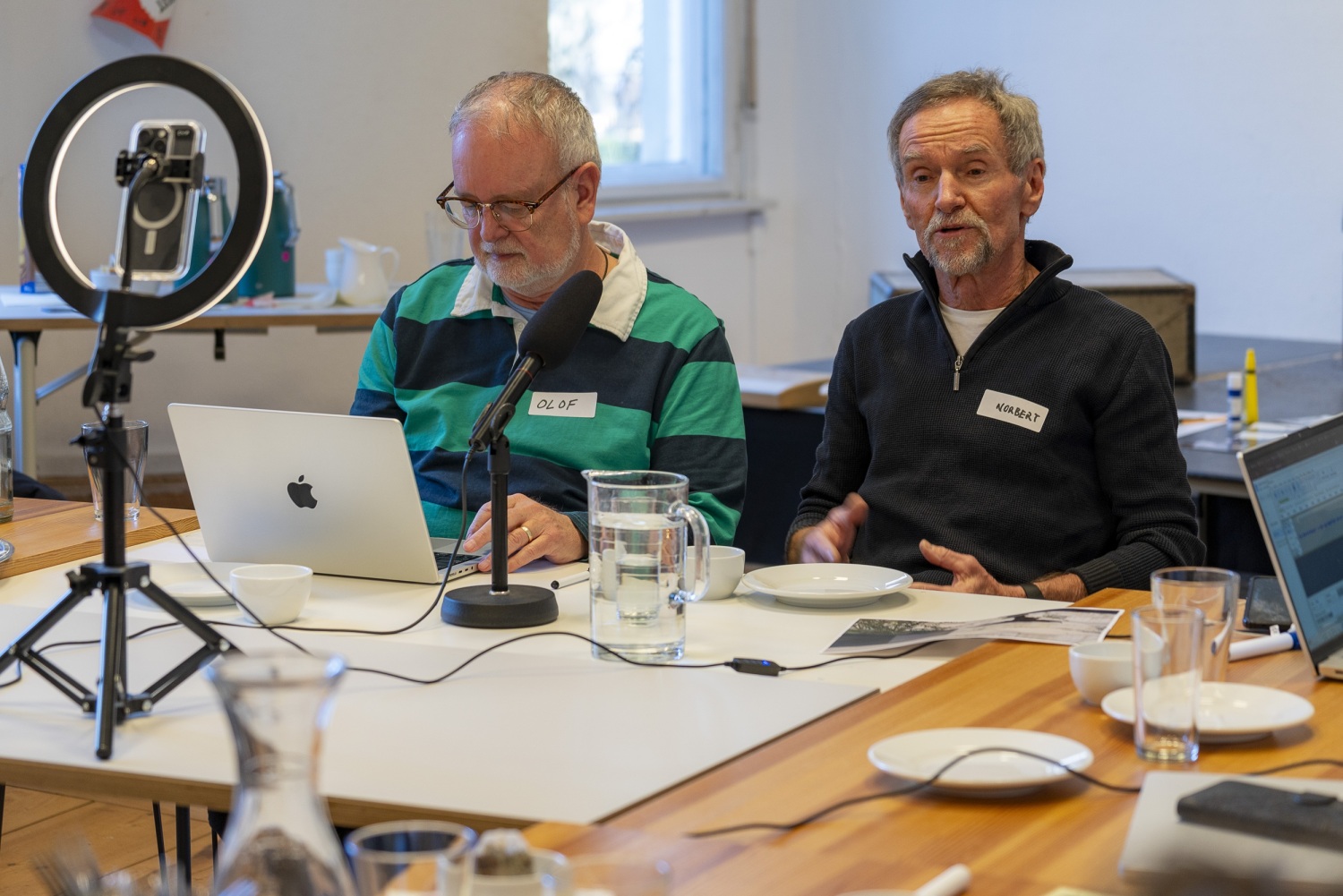 Zwei Männer sitzen nebeneinander an einem Tisch. In der Mitte steht ein Mikrofon.