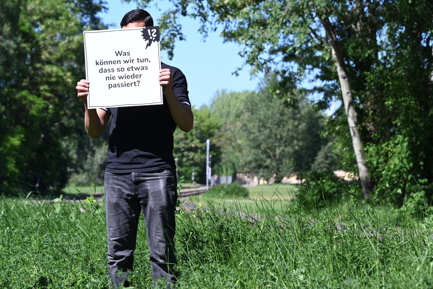 Jemand steht auf einer Grünfläche und hält ein Schild in den Händen, auf dem steht, "Was können wir tun, dass so etwas nie wieder passiert?"