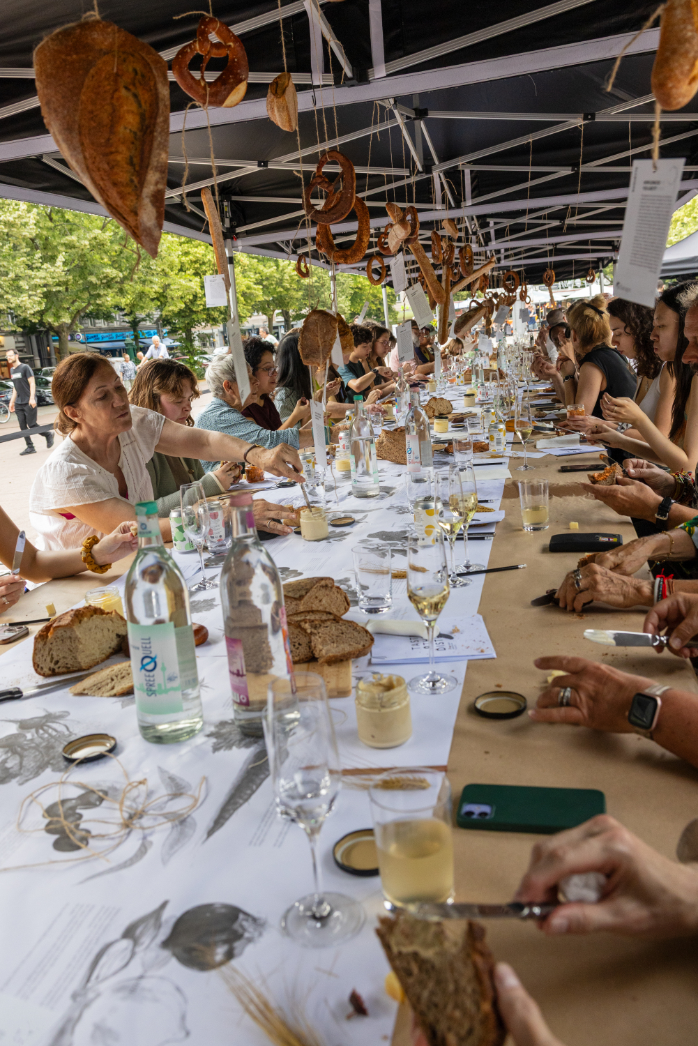 Viele Menschen sitzen an einer langen Tafel überdacht von einem Pavillon von welchem verschiedenste Brote herab hängen. Die Brote reichen von Brezeln, über Sauberteigbrot, bis hin zum Simit (Sesamring) oder Baguettes. Die Tafel ist mit einer schönen Tischdecke verziert. Die Menschen bedienen sich an den verschiedenen Broten und Dips, welche in der Mitte der Tafel stehen, selbst.