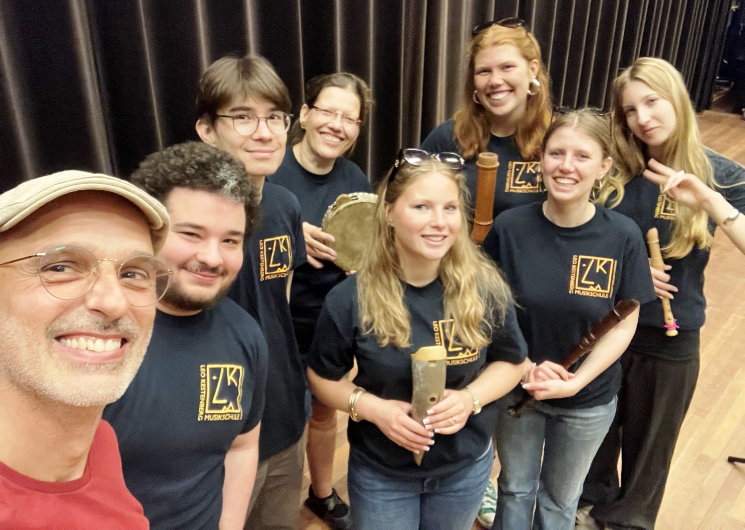 Sieben Personen mit T-Shirts der Leo Kestenberg Musikschule stehen mit Instrumenten zusammen. Im Vordergrund lächelt ein weiterer Mann in die Kamera.