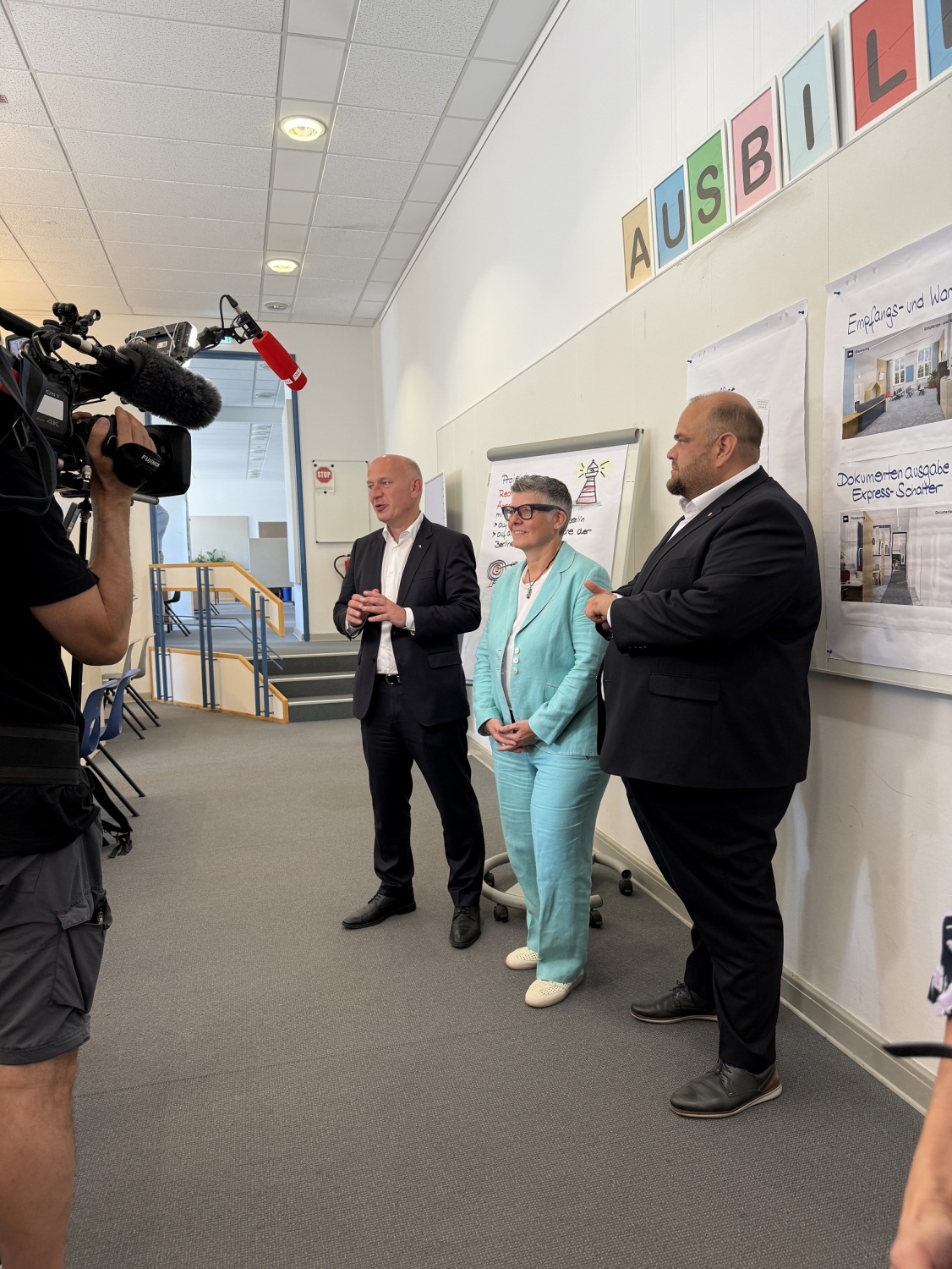 Drei Personen stehen vor einer Wand mit Infomaterial und einem Flipchart. Sie werden von einem Kamerateam aufgenommen.