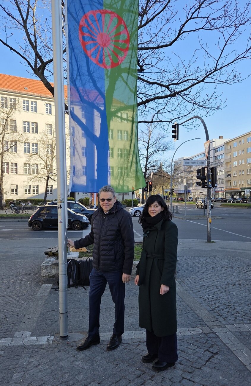 Zwei Personen stehen neben einem Fahnenmast, an dem eine Flagge mit rotem Rad auf blau-grünen Hintergrund abgebildet ist.