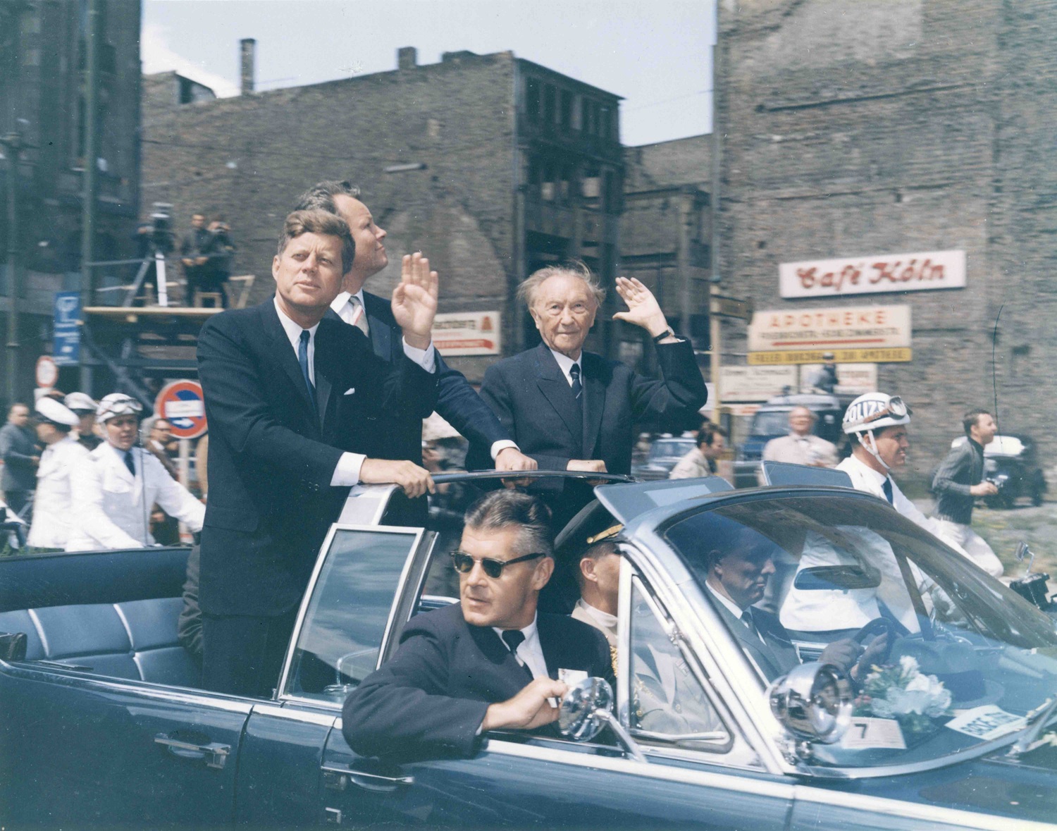 26. Juni 1963 Die ursprüngliche Bildunterschrift auf der Rückseite des Abzugs lautet: „Präsident John F. Kennedy am ‚Checkpoint Charlie‘ mit dem West-Berliner Bürgermeister William Brandt und dem westdeutschen Bundeskanzler Konrad Adenauer [der winkt].“ Berlin, Deutschland