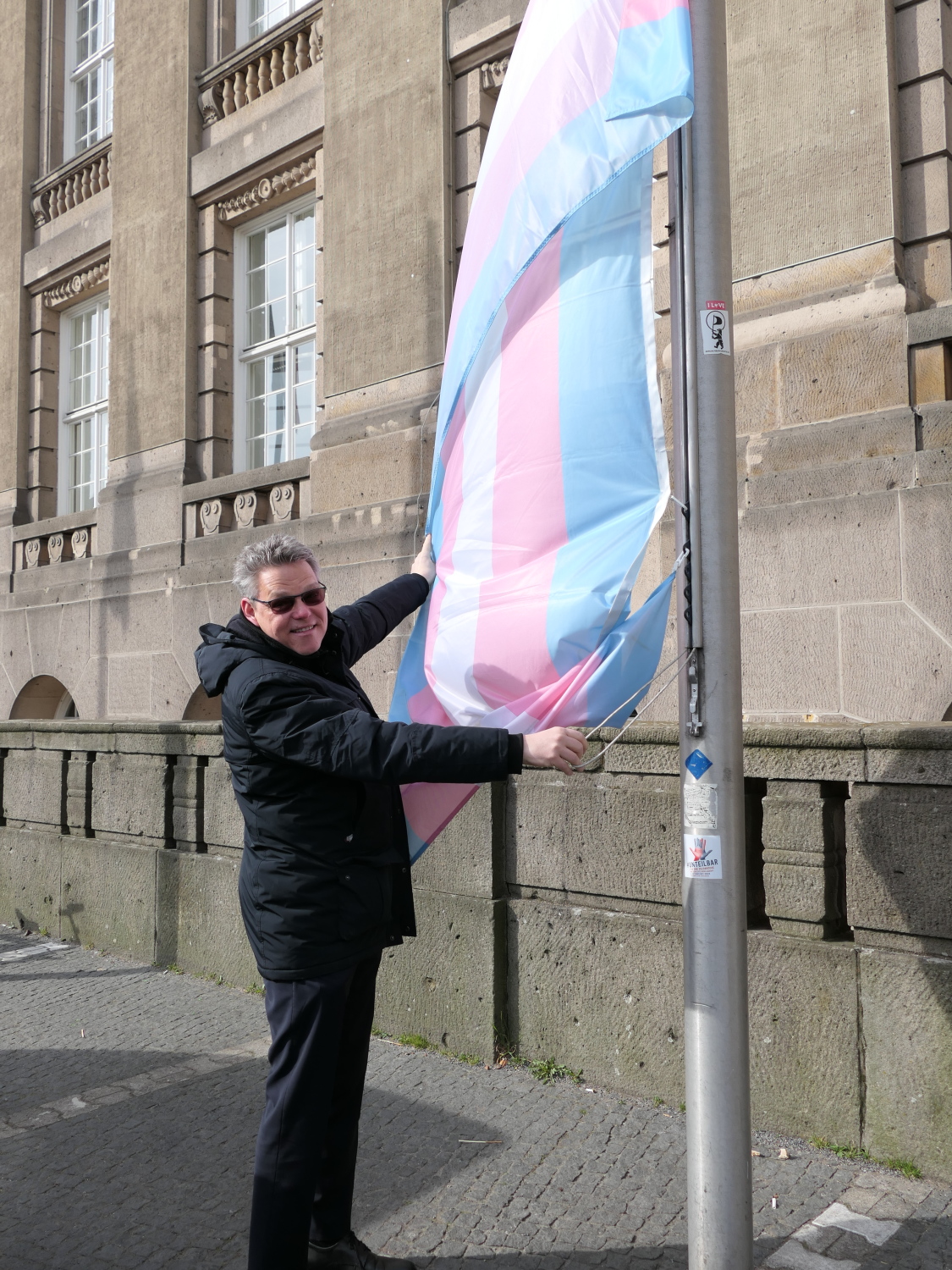 Ein Mann steht neben einem Fahnenmast und hält eine an diesem hängenden blau-rosa-weiß-gestreifte Flagge in den Händen.