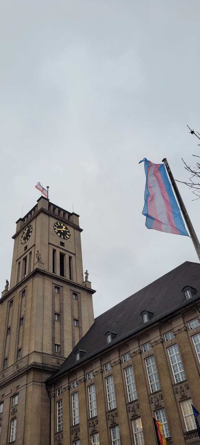 Vor einem großen Gebäude mit Uhrenturm weht eine blau-rosa-weiß-gestreifte Flagge an einem Mast.