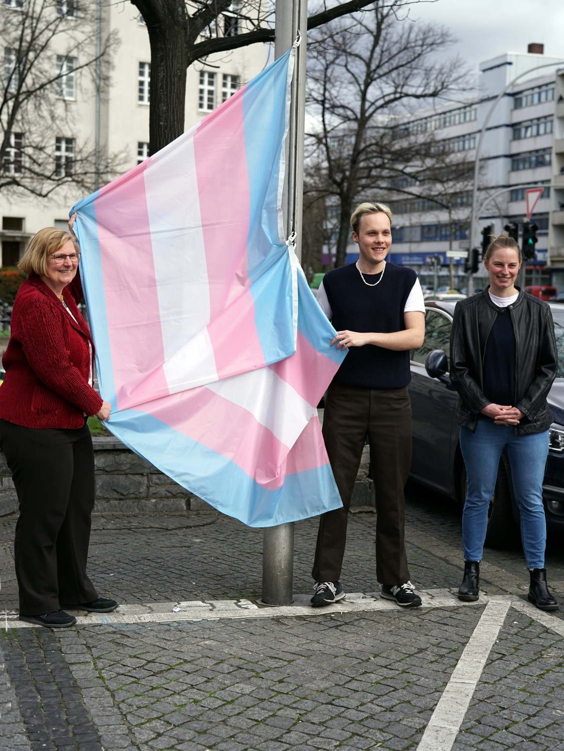 Zwei Frauen und ein Mann stehen neben einem Fahnenmast und halten eine blau-rosa-weiß-gestreifte Flagge.
