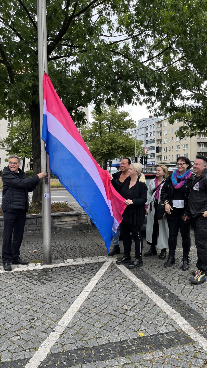 Eine Gruppe Menschen steht bei einem Fahnenmast. Ein Mann hält an dem Fahnenmast eine pink-lila-blau-gestreifte Flagge hoch.