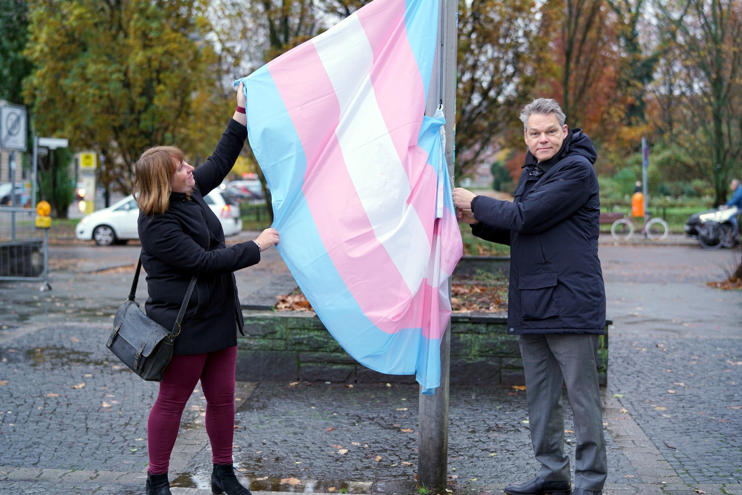 Eine Frau und ein Mann stehen an einem Fahnenmast und halten eine blau-rosa-weiß-gestreifte Fahne hoch.