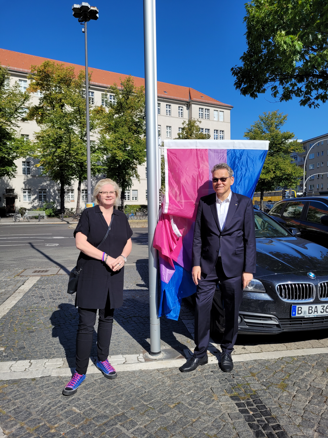 Eine Frau und ein Mann stehen vor einem Fahnenmast, an dem eine pink-lila-blaue Flagge hängt.