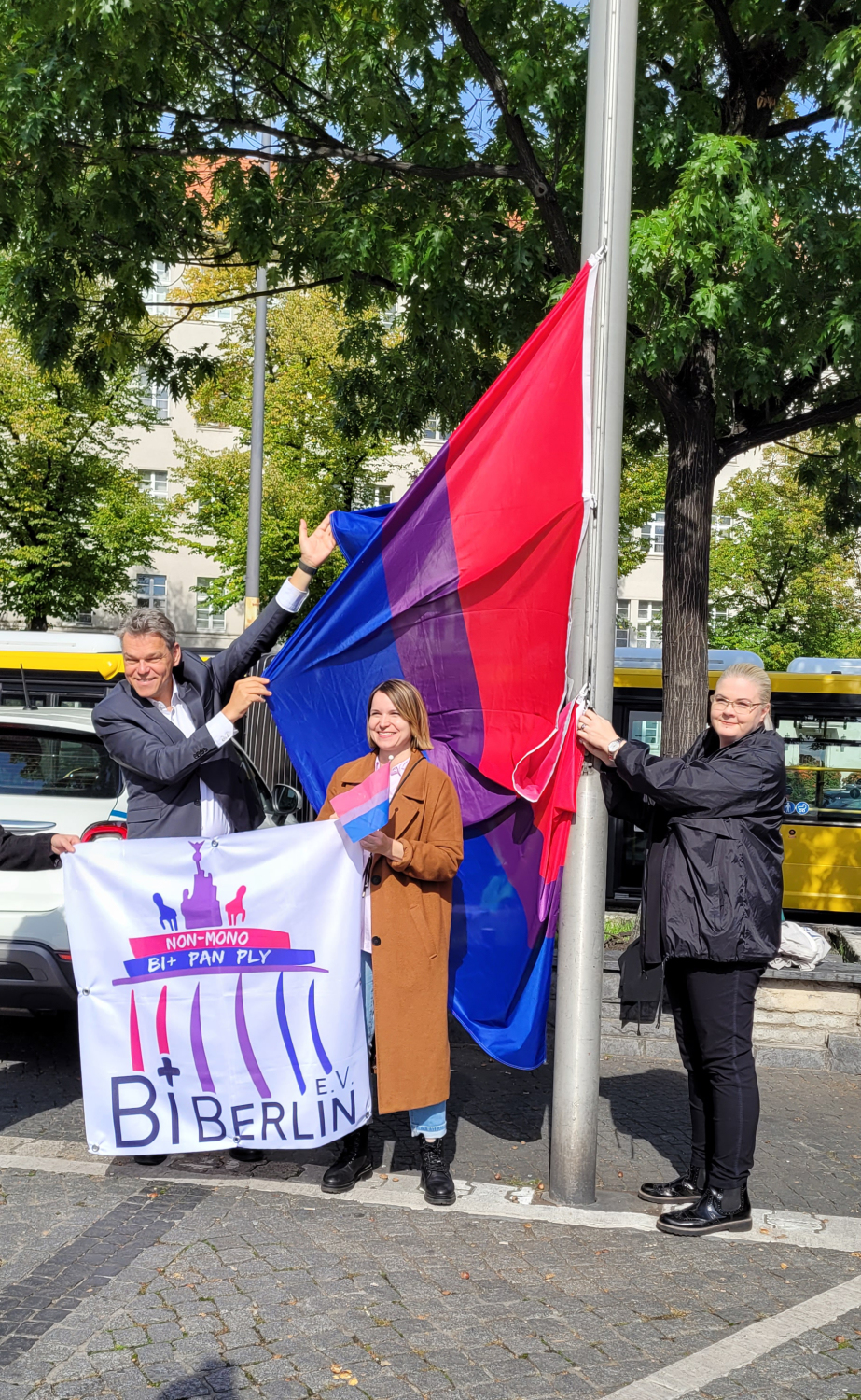 Ein Mann und zwei Frauen an einem Fahnenmast halten eine blau-lila-pink-gestreifte Flagge und eine kleine Flagge mit dem Logo von BiBerlin.
