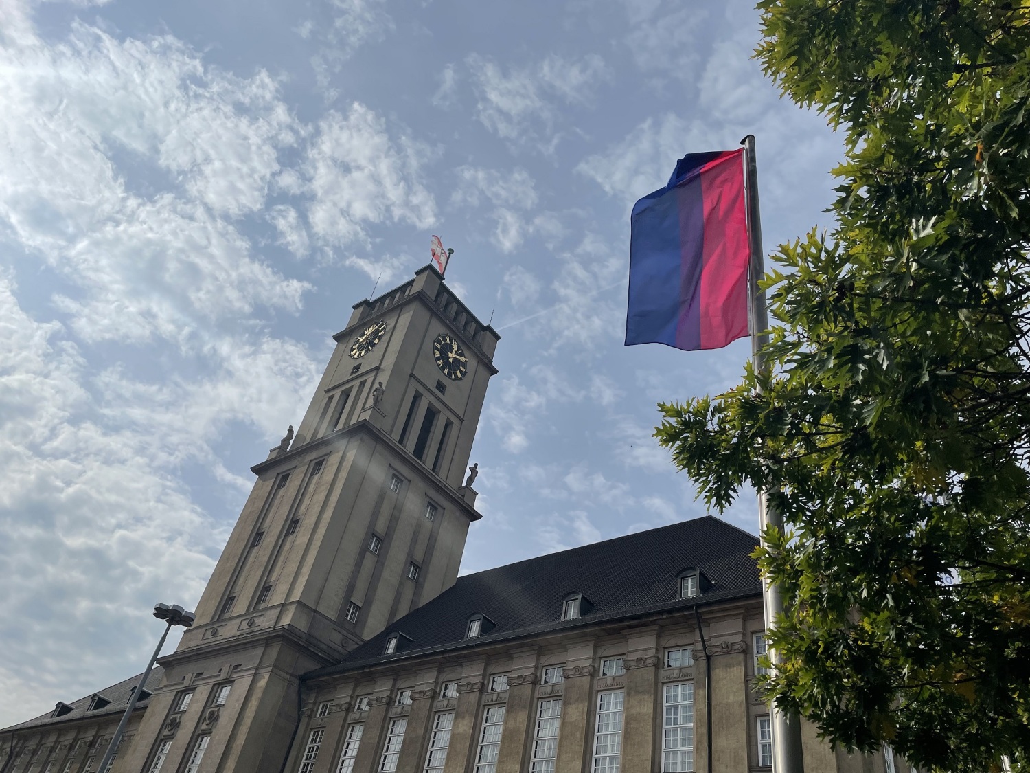 Vor einem großen Gebäude mit Uhrenturm weht eine blau-lila-pink-gestreifte Flagge an einem Fahnenmast.