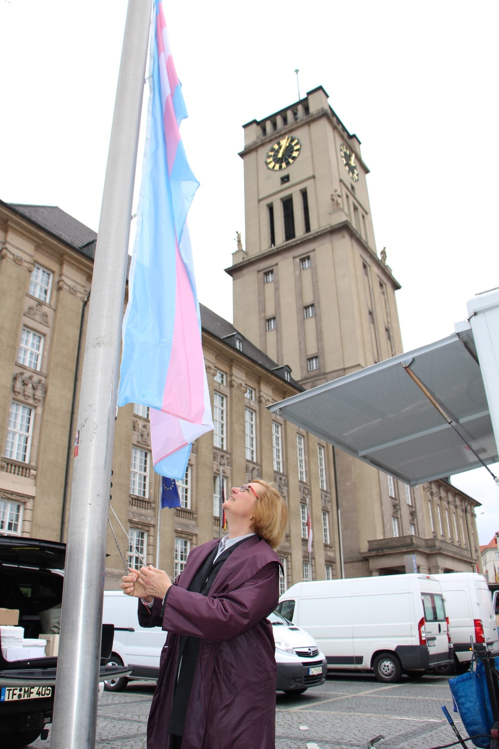 Eine Frau hisst vor einem großen Gebäude mit Uhrenturm eine blau-rosa-weiß-gestreifte Flagge.