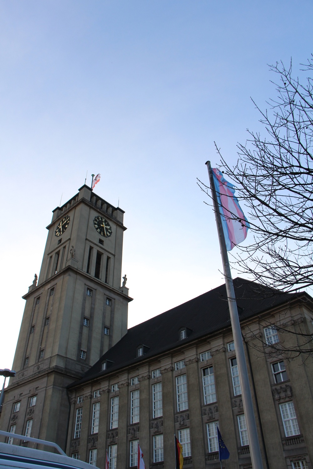 Vor einem großen Gebäude mit Uhrenturm weht eine blau-rosa-weiß-gestreifte Flagge an einem Mast.