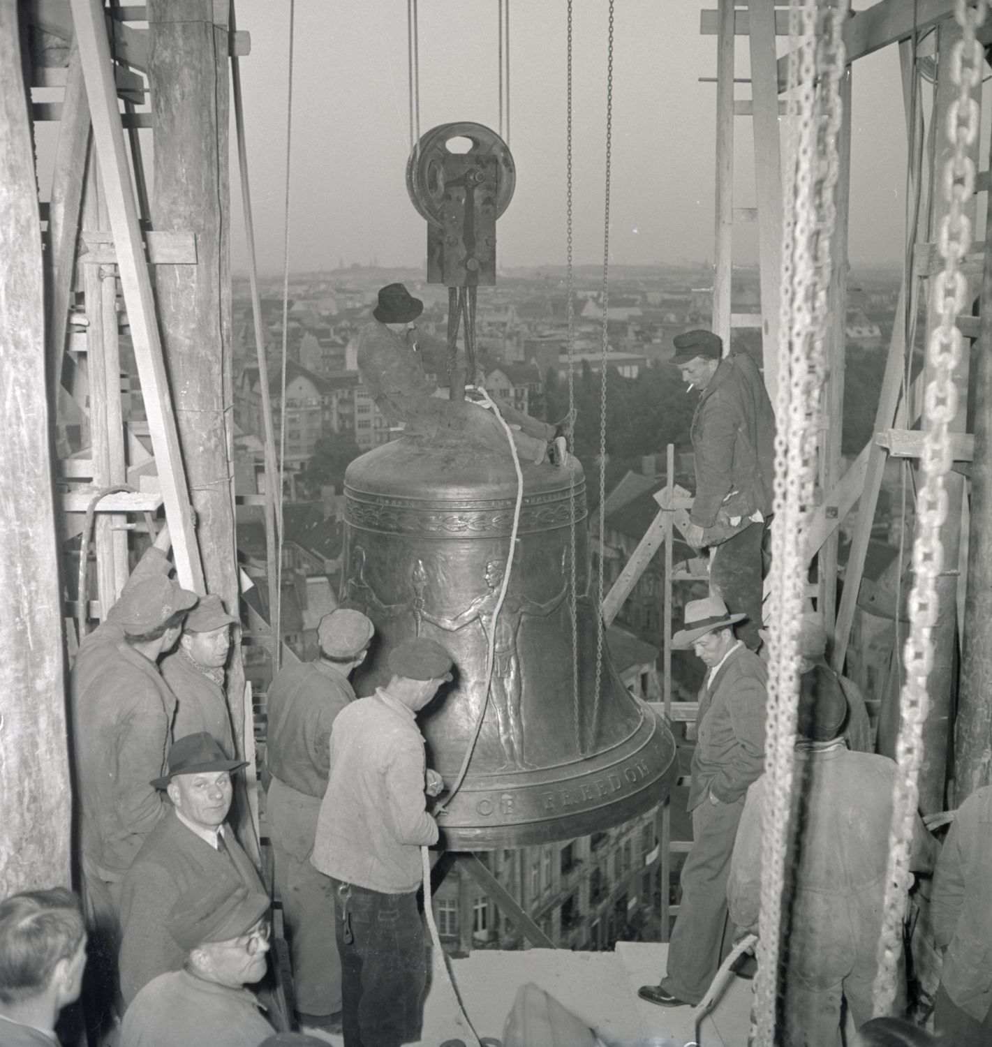 Several men are pulling a large bell into a bellfry using a winch. In the background are the roofs of a big city.