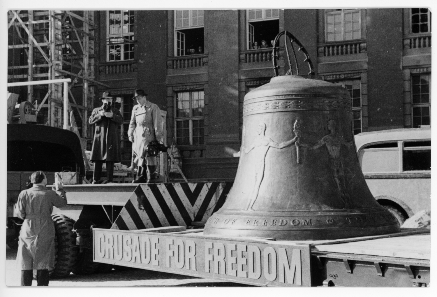 A large bell is being transported on a low loader, on the side is the inscription "Crusade for Freedom". Three men are standing near the bell.