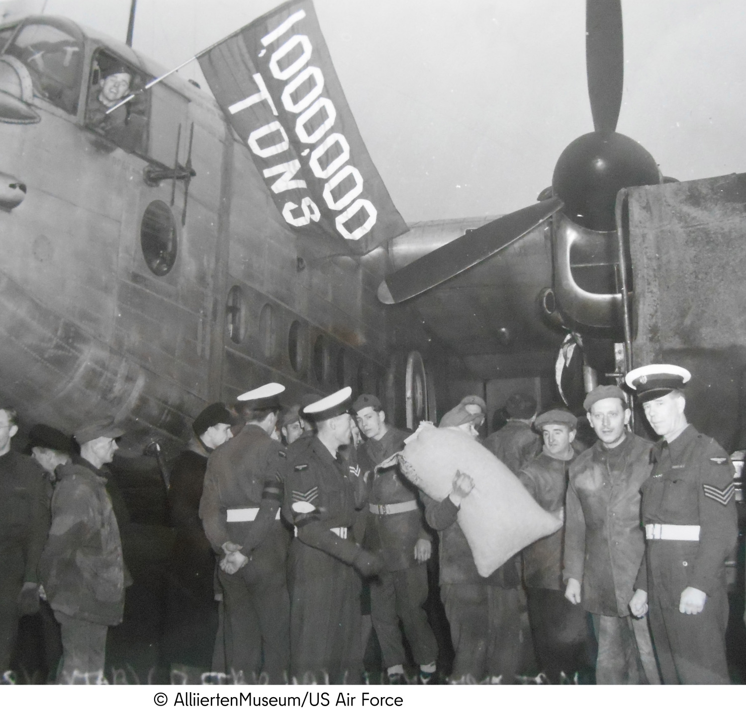 Various men in uniform stand in front of an airplane and carry sacks of groceries