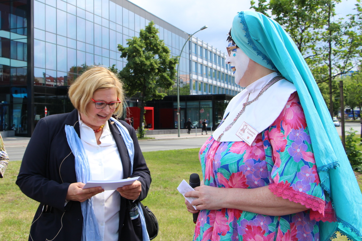 Links eine Frau schaut auf ihre Notizen, rechts eine bunt gekleidete Frau mit einem türkisen Kopftuch wie bei einer Nonne mit weiß bemaltem Gesicht