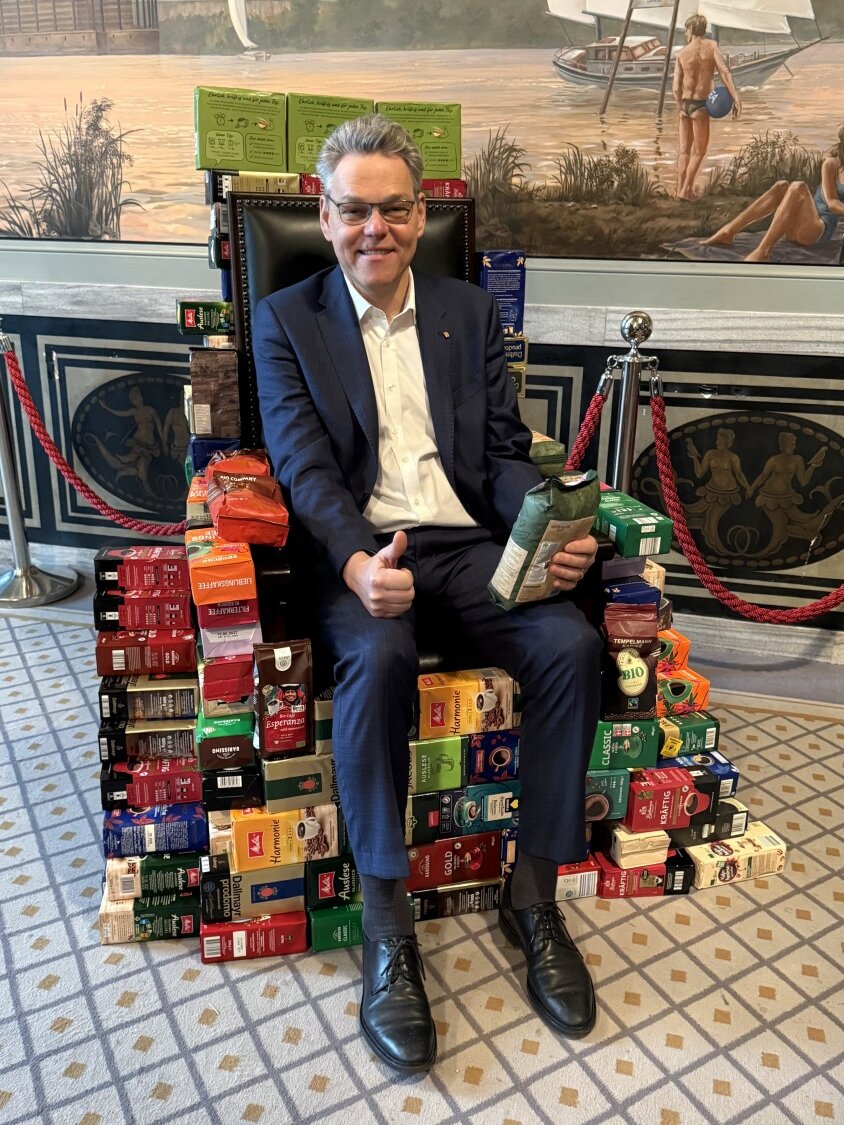Bezirksbürgermeister Jörn Oltmann sitzt auf einem Stuhl, der durch diverse Kaffeepakete umrahmt wird.