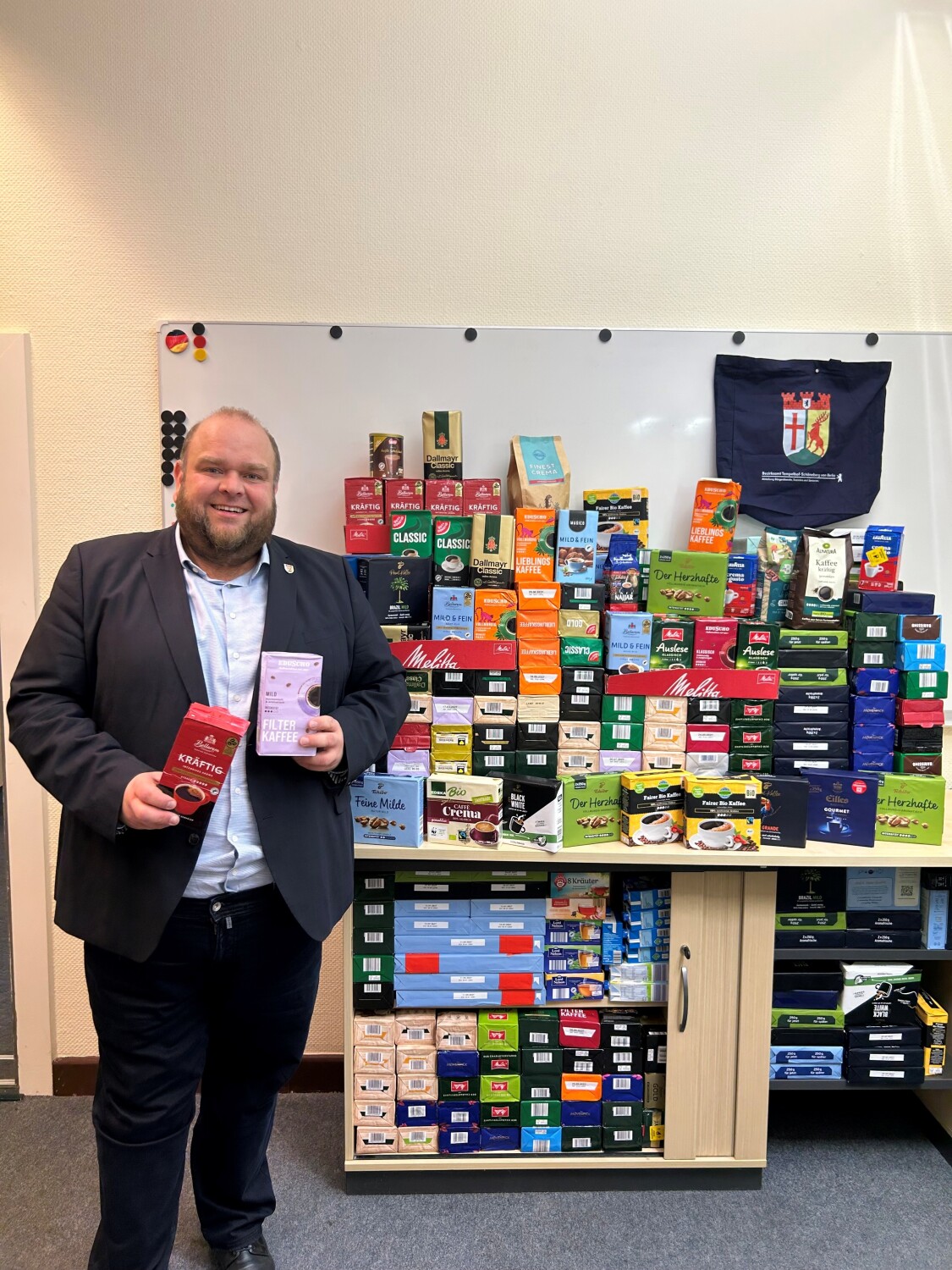 Stellvertretender Bezirksbürgermeister und Bezirksstadtrat Matthias Steuckardt hält zwei Kaffepakete in seinen Händen und steht vor einem Tisch, auf dem diverse Kaffepakete gestapelt sind.
