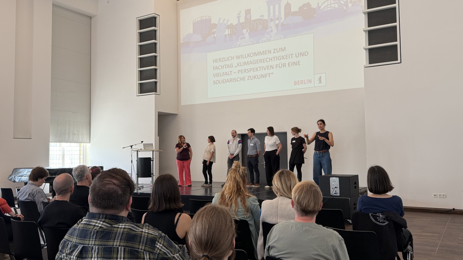 Sieben Personen stehen vor Publikum auf einer Bühne eines großen Saales. An die Wand ist eine Präsentation projiziert.