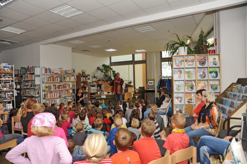 Ein Märchenerzähler spricht vor einer großen Anzahl Kinder und erzählt Märchen . Die Kinder sitzen auf Kissen und Stühlen und hören ganz begeistert zu.