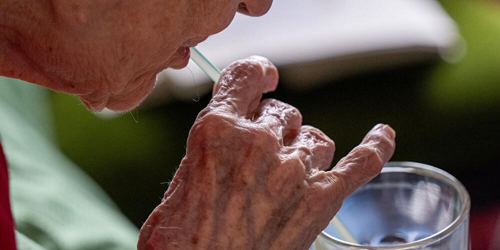 Nahaufnahme einer älteren Person, die mit einem Strohhalm aus einem Glas trinkt.