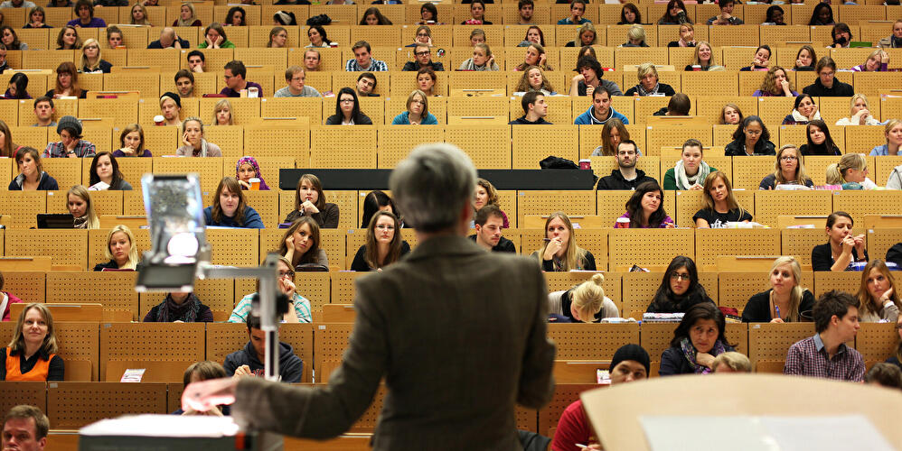 Vorlesung in einem großen Hörsaal: Eine Professorin spricht vor zahlreichen Studierenden auf ansteigenden Sitzreihen.