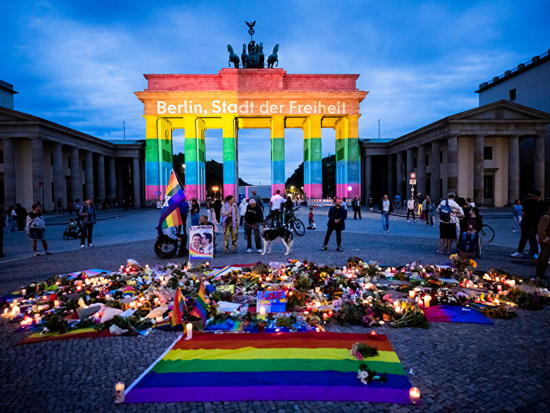 Brandenburger Tor in Regenbogenfarben, davor Kerzen, Blumen und Pride-Flaggen