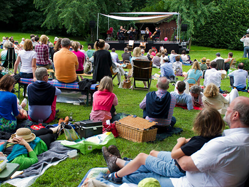 Besucher:innen sitzen auf einer Wiese vor einer Bühne und hören Musik.