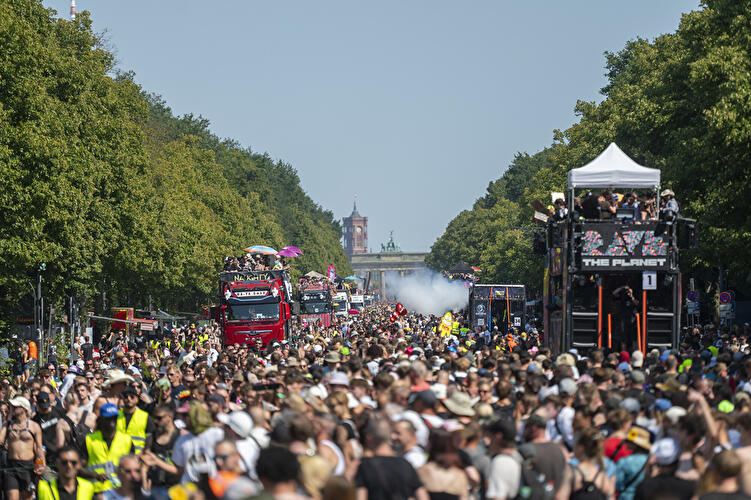 Dichte Menschenmenge auf der Straße des 17. Juni bei der Rave the Planet Parade in Berlin, umgeben von Bäumen, mit Musik-Trucks und Blick auf das Brandenburger Tor im Hintergrund.